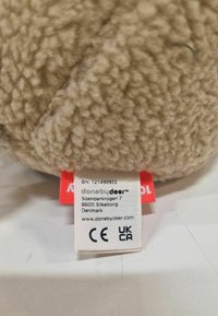 Beež pehme mänguasi, millel on tekstureeritud, karvane pind. Omab valget silti punaste aktsentidega, kus on näidatud bränd ja ohutuse sertifikaadid.