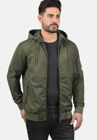 Veste à capuche verte olive en tissu lisse avec une fermeture éclair à l'avant, des poignets côtelés et des poches latérales ; comprend une poche zippée sur la manche.