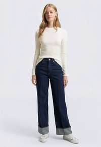 Haut à manches longues beige clair associé à un jean taille haute bleu foncé à jambes larges avec un ourlet retroussé, porté avec des baskets blanches.