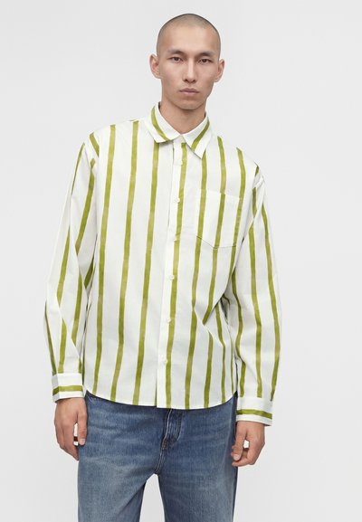 Άνδρας που φοράει λευκό πουκάμισο με κάθετες ελαιίστικες πράσινες ρίγες και μπλε τζιν, στέκεται μπροστά σε απλό ανοιχτό γκρι φόντο.