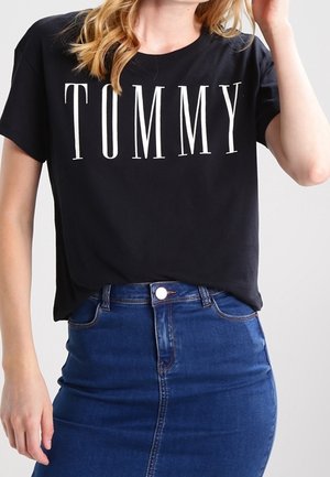 Vrouw die een zwart T-shirt draagt met de tekst "TOMMY" en een blauwe spijkerrok, poseert met één hand bij haar hoofd en de andere ontspannen.