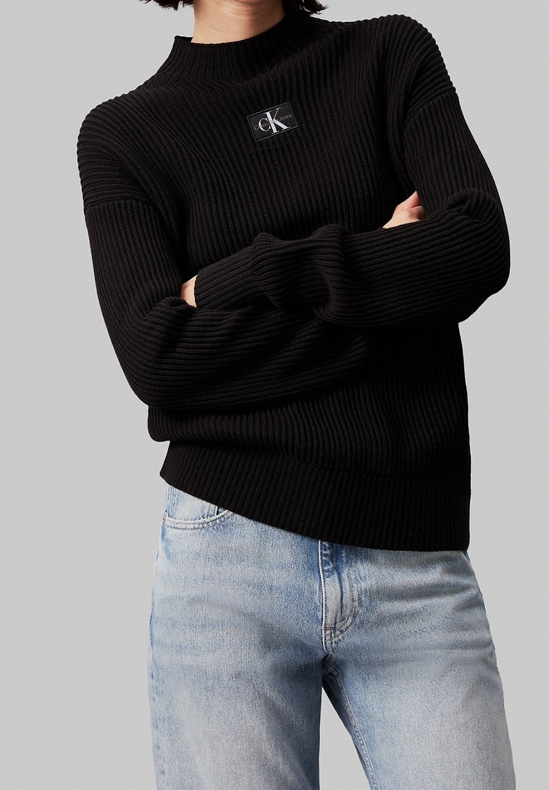 Personne portant un pull noir côtelé avec un patch logo Calvin Klein et un jean bleu clair, les bras croisés, devant un fond gris uni.