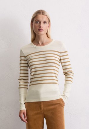 Femme blonde portant un haut à rayures horizontales beiges et brunes à manches longues avec un pantalon brun, se tenant contre un mur blanc texturé.