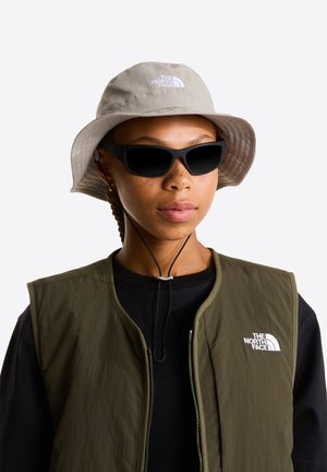 Sombrero tipo cubo gris claro con logo, gafas de sol negras y chaqueta sin mangas verde oliva. Texturas suaves y diseño práctico.