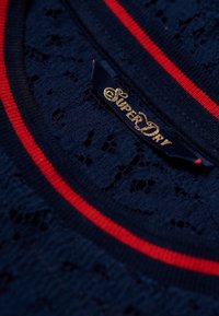 Mörkblått stickat tyg med spetsstruktur, med rund hals och röda randiga detaljer. "SuperDry"-etikett i guld på en mörk bakgrund.