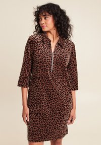 Vrouw in een bruine jurk met luipaardprint met driekwartmouwen en een ritssluiting aan de voorkant, kijkend naar links tegen een beige achtergrond.