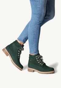Gröna ankelstövlar i mocka, med metallögon och tjocka snören, med en kraftig brun sula, visade tillsammans med blå jeans.