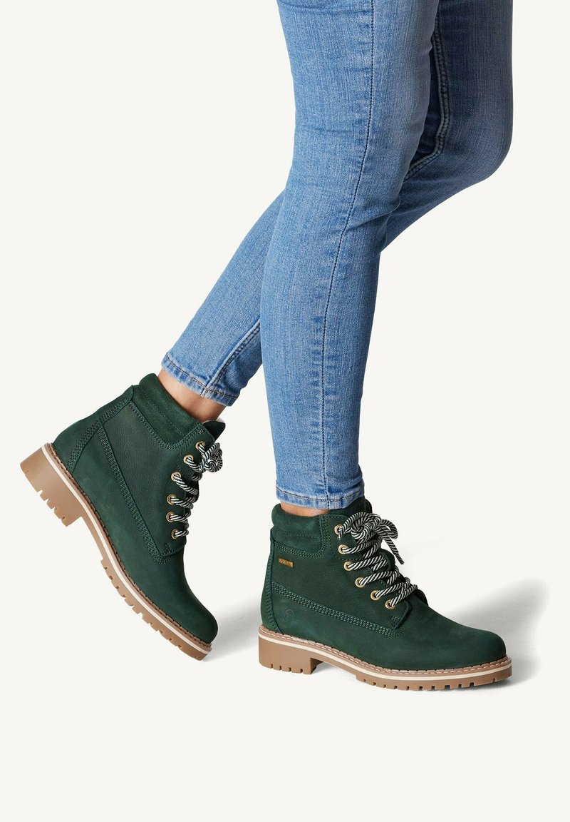 Gröna ankelstövlar i mocka, med metallögon och tjocka snören, med en kraftig brun sula, visade tillsammans med blå jeans.