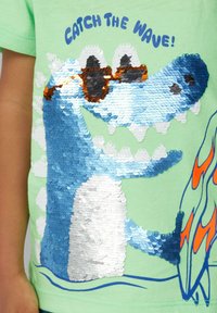 Groene katoenen T-shirt met een met pailletten versierde blauwe alligator met zonnebril, golfpatroon en tekst "CATCH THE WAVE!" in het blauw.