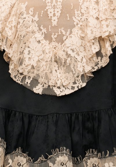 Μπεζ υφασμάτινη φλοραλ δαντέλα στρωμένη πάνω σε μαύρο ύφασμα με ρεβέρ στο τελείωμα και λεπτομέρειες δαντέλας στο κάτω άκρο.