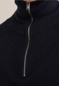 Detailní záběr na tmavě modrý svetr s vysokým límcem a polo zipem, s texturovaným pleteným vzorem a stříbrným detailem na zipu.