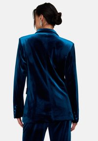 Samtblazer in tiefem Petrol, taillierte Passform mit Reverskragen, einzelner Rückennaht und geknöpften Manschetten. Glatte Textur und sichtbarer Glanz.