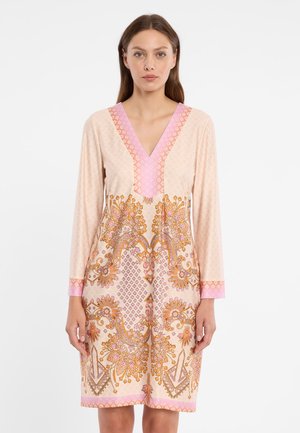 Frau trägt ein langärmliges, knielanges Kleid mit rosa und orangefarbenen Blumen- und geometrischen Mustern auf beigefarbenem Hintergrund.