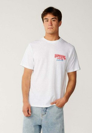 Jeune homme portant un t-shirt blanc avec un texte rouge et bleu sur la poitrine et un jean bleu clair, debout devant un fond uni.