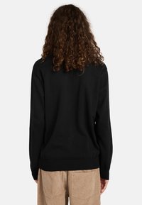 Maglione nero con maniche lunghe e fondo a coste, realizzato in un materiale morbido. Indossato sopra pantaloni in velluto a coste color marrone chiaro, presenta un design semplice.