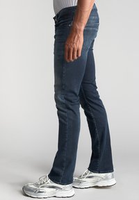 Jeans en denim bleu foncé avec une coupe slim, un design à cinq poches et un léger délavage. Portés avec des chaussures de sport argentées et un t-shirt blanc.