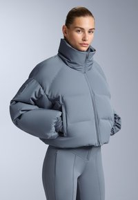 Sivi puffer žaket s visokim ovratnikom, s prednjim patentnim zatvaračem i voluminoznim, podstavljenim dizajnom. Glatka tekstura i prilagođeni kroj.