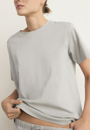 Femme portant un t-shirt à manches courtes gris clair et un pantalon à taille élastique, soulevant légèrement le t-shirt pour dévoiler la taille.