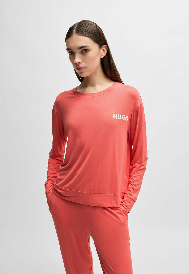 Korall långärmad topp med rund halsringning, prydd med "HUGO"-logotyp i vitt. Tillverkad av ett slätt, elastiskt tyg med subtil textur.