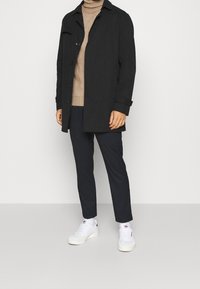 Cappotto nero con bottoni sopra un dolcevita beige, abbinato a pantaloni blu marino e sneakers bianche. Linee pulite e design minimalista in tutto.