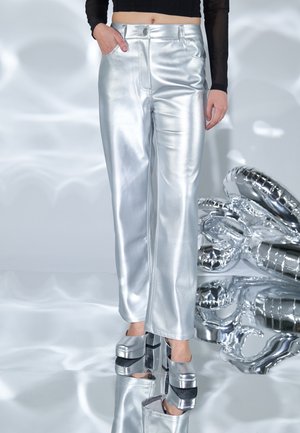 Person wearing shiny silver pants and matching platform heels, standing on reflective surface next to metallic silver balloon arrangement.