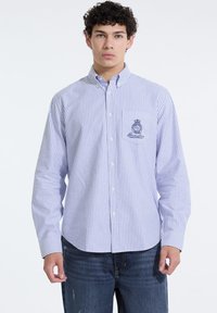 Camisa de botones a rayas en azul claro y blanco, con un bolsillo parchado y un logo bordado en el pecho. Fabricada en tejido de algodón entrelazado.