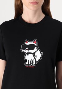 Černé bavlněné tričko s potiskem karikatury bílé kočky s červenými akcenty a slunečními brýlemi, umístěné ve středu, s textem "KARL LAGERFELD" pod ním.