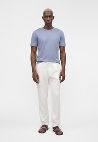 Homme aux cheveux blonds courts portant une chemise bleu clair à manches courtes, un pantalon blanc à cordon et des sandales marron foncé, debout sur un fond blanc.