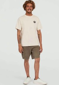 Smiling man with curly hair wearing a beige t-shirt, brown cargo shorts, white sneakers, and visible arm tattoos, standing against a plain background.