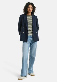 Marineblå nålestribet blazer med guldknapper, parret med en grøn og hvid stribet skjorte, lyseblå jeans og beige sneakers.