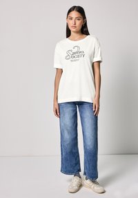 Weißes Baumwoll-T-Shirt mit schwarzem Grafiktext, kombiniert mit hellblauen weit geschnittenen Jeans. Das Model trägt beiges Sneaker mit einer glatten Textur.