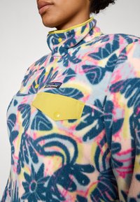 Fleece-Pullover im lebhaften, mehrfarbigen Blumenmuster, mit hohem Kragen, gelbem Druckknopfverschluss und einer Brusttasche mit gelbem Akzent.