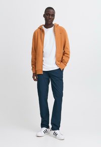 Orange zip-up hoodie med dragsko i huven, kombinerad med en vit t-shirt och marinblå byxor. Skor inkluderar vita sneakers med svarta ränder.