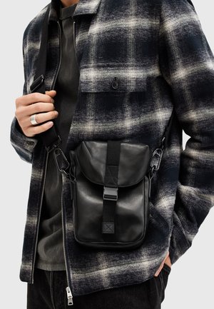 Homme portant une veste à carreaux noirs et blancs tenant un sac bandoulière en cuir noir avec une boucle.