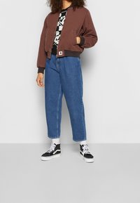 Hnědá bombardovací bunda s žebrovanými manžetami, černé tričko s grafikou, džíny s vysokým pasem modré barvy a černé tenisky s bílými akcenty.