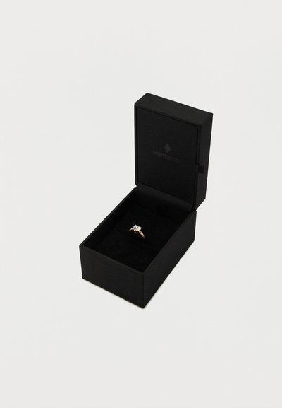 Kuldne sõrmus südamekujulise kiviga, esitatud mustas karbis sametise sisevoodriga. Karbi kaanel minimalistlik logo.