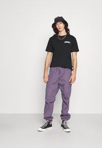 T-shirt en coton noir avec logo blanc, pantalon cargo violet avec taille élastique, et baskets montantes noires avec accents blancs.