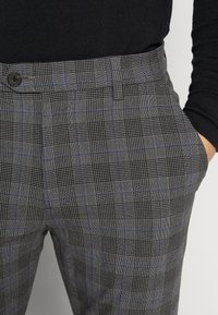 Pantalon à carreaux gris avec un motif bleu subtil, doté d'une fermeture à boutons, de passants pour ceinture et d'une coupe ajustée. Détail du tissu texturé visible.