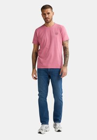 Rosa Baumwoll-T-Shirt mit rundem Ausschnitt und kleinem Grafikdruck auf der linken Brust. Kombiniert mit blauen Denim-Jeans und weißen Sneakers.