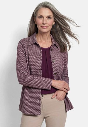 Reife Frau mit langen grauen Haaren trägt eine lilafarbene gemusterte Jacke über einem dunkelvioletten Oberteil und beigen Hosen, steht mit einer Hand in der Tasche.