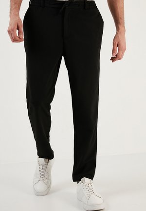 Pantalons noirs à taille élastique avec une coupe décontractée et cordon de serrage. Portés avec des baskets blanches, présentant un tissu lisse et des poches latérales.