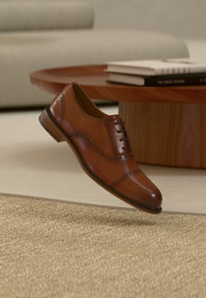 Brauner Brogue-Lederschnürschuh schwebt über einem beigen Teppich, im Hintergrund ein runder Couchtisch aus Holz und gestapelte Bücher.