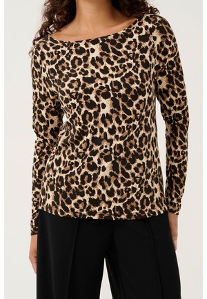 Vrouw draagt een lange mouwen top met luipaardprint, gecombineerd met zwarte broek, gefotografeerd van nek tot heupen tegen een neutrale achtergrond.