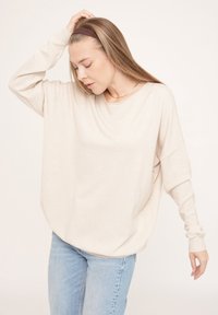 Beige übergroßes Langarmoberteil aus weichem Stoff mit rundem Ausschnitt und gesenkten Schultern, kombiniert mit hellblauen Jeans.
