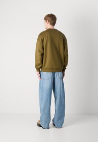 Felpa verde oliva con collo rotondo e polsini a costine, abbinata a jeans a gamba larga blu chiaro. Il modello è girato di spalle, mostrando i dettagli posteriori.