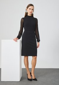 Black dress with fitted bodice, high neckline, and long sheer polka-dotted sleeves. Smooth texture, knee-length, paired with black high heels.