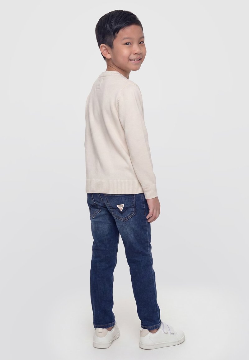 Garçon souriant par-dessus son épaule, portant un pull crème, un jean bleu et des baskets blanches, debout devant un fond clair uni.