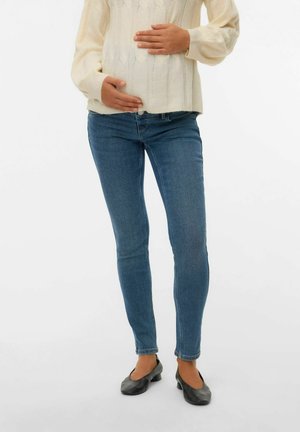 Jean skinny bleu avec une texture lisse, associés à un pull en tricot torsadé crème. Le modèle berce un ventre bien visible. Chaussures plates noires.