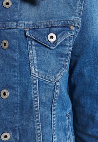 Niebieska kurtka dżinsowa z srebrnymi elementami, z kieszenią na piersi, zapięciem na guziki oraz widocznymi detalami ściegów. Teksturowany materiał.