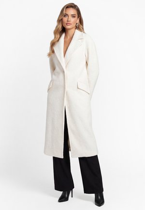 Donna che indossa un lungo cappotto bianco testurizzato sopra pantaloni neri e stivali neri con tacco, in piedi con le mani nelle tasche del cappotto su sfondo bianco.